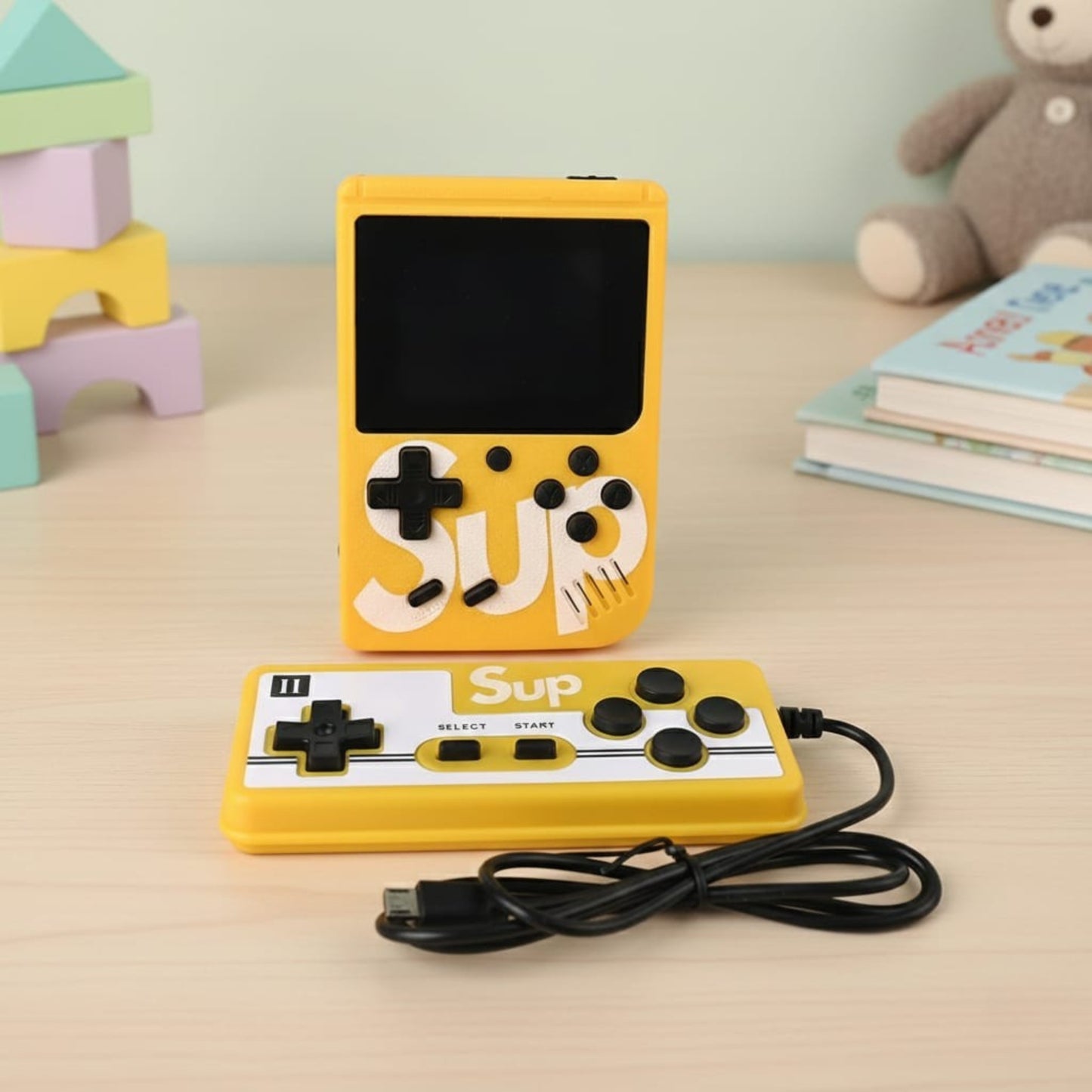Yellow handheld gaming console with 'SUP' branding on a table with toys and books in the background - Baby Nest